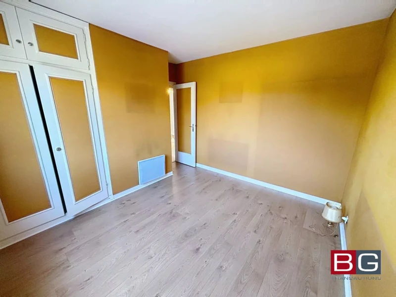 La photo montre une pièce vide avec des murs jaunes, un sol en bois clair, une porte blanche et un placard intégré.