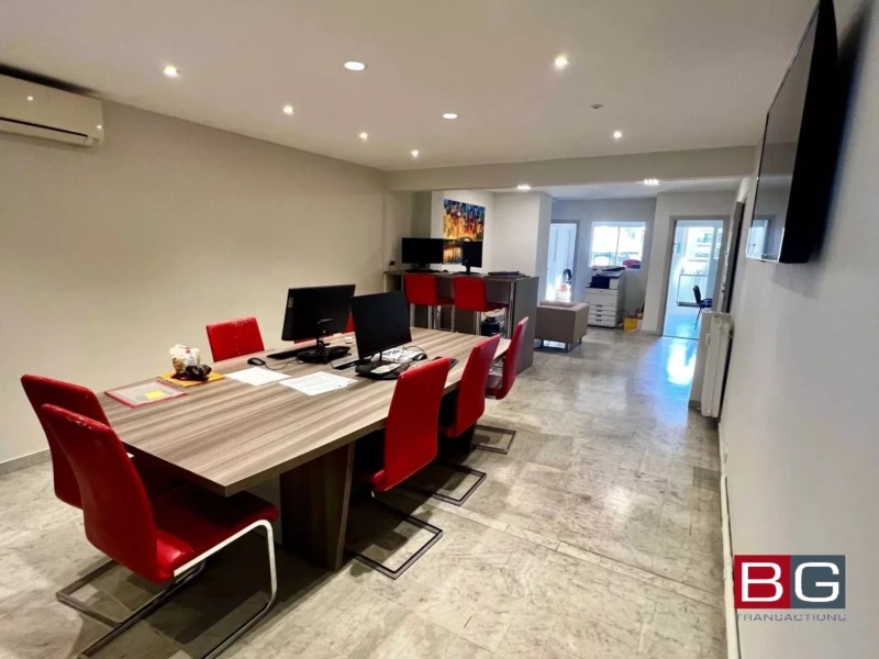 C'est un bureau moderne avec des chaises rouges autour d'une grande table et des ordinateurs installés. L'éclairage est encastré au plafond, et l'espace est bien organisé.