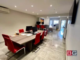 C'est un bureau moderne avec des chaises rouges autour d'une grande table et des ordinateurs installés. L'éclairage est encastré au plafond, et l'espace est bien organisé.