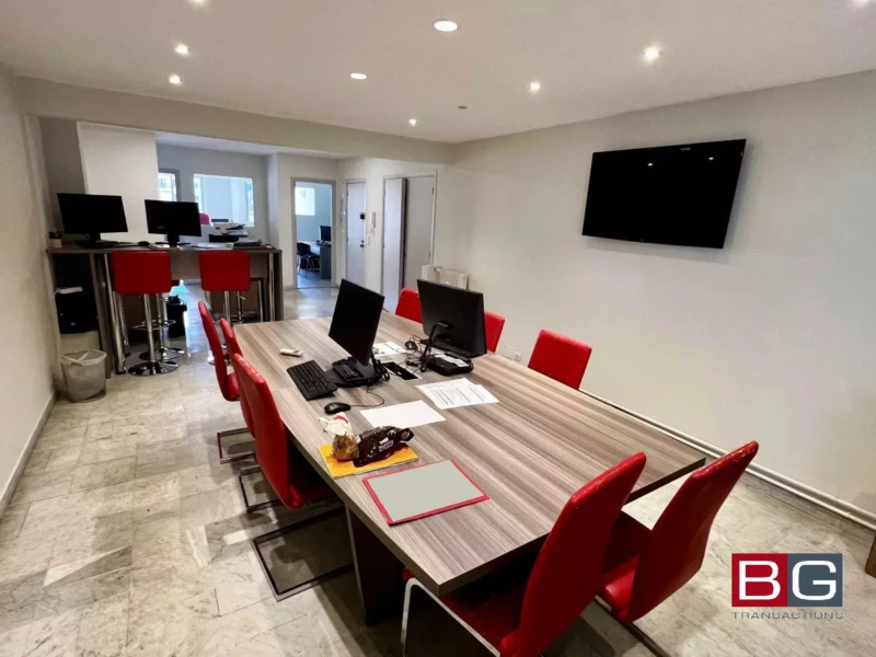 Une salle de réunion moderne avec une grande table entourée de chaises rouges et des ordinateurs sur le bureau. Il y a un écran fixé au mur et un espace de travail visible en arrière-plan.