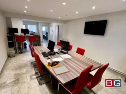 Une salle de réunion moderne avec une grande table entourée de chaises rouges et des ordinateurs sur le bureau. Il y a un écran fixé au mur et un espace de travail visible en arrière-plan.