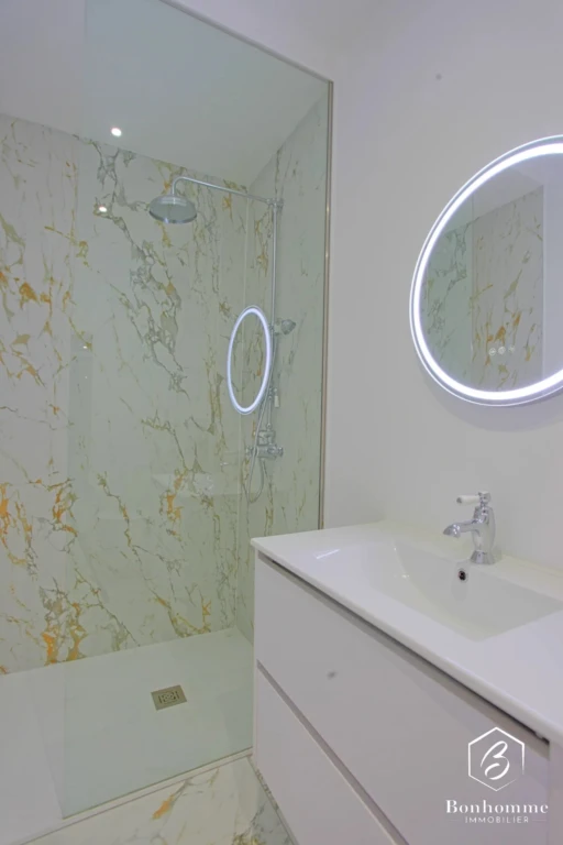 Salle de bain moderne avec douche à l'italienne et lavabo, ornée de marbre et éclairée par un miroir rond rétroéclairé.