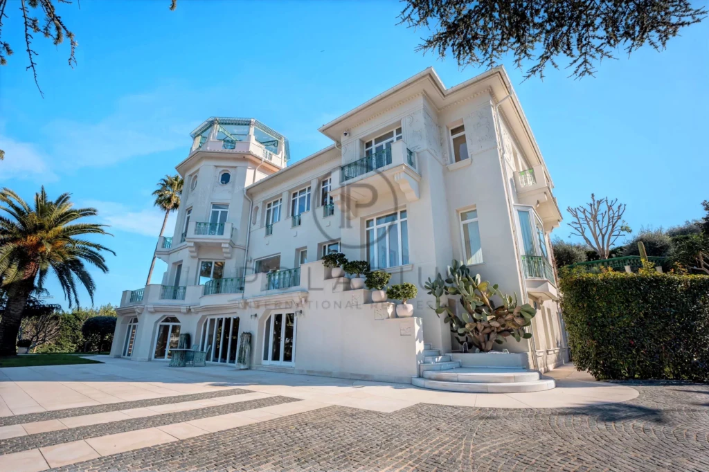 Grande villa blanche avec plusieurs balcons et palmiers, entourée de verdure sous un ciel bleu.