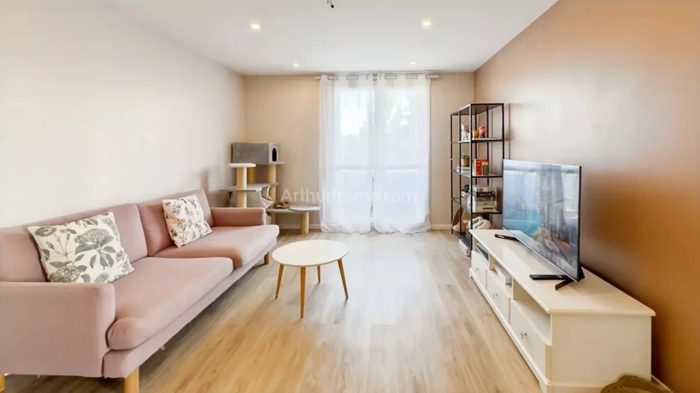 PONTHIERRY F4 Un salon moderne avec un canapé rose, une table basse ronde, et une étagère à côté d'une télévision. Les murs sont peints en marron et blanc, avec un sol en bois clair.