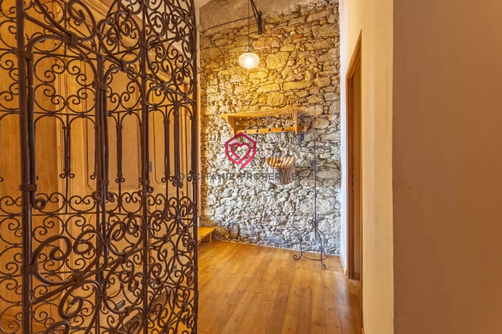 Un couloir chaleureux avec un mur en pierre apparente, un sol en bois, et une porte ornée de fer forgé.