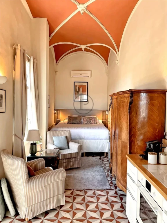 Une chambre élégante avec un plafond voûté, un lit double, un fauteuil, une armoire en bois, et un sol à motifs géométriques.
