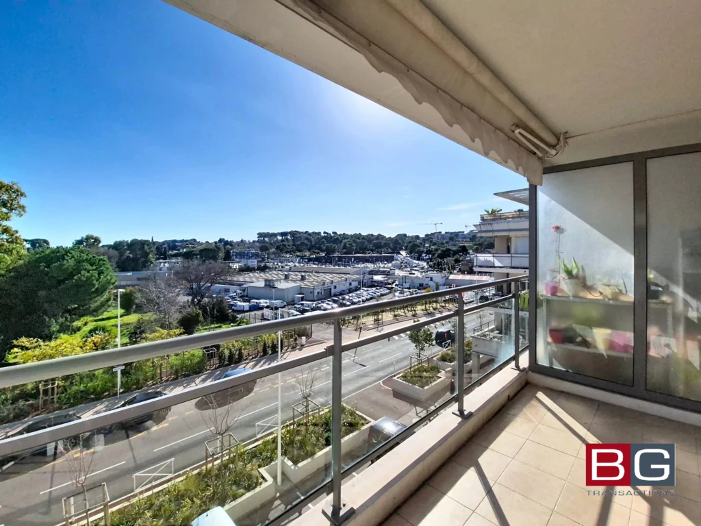 Balcon ensoleillé avec vue sur la route, la verdure et un port de plaisance au loin.