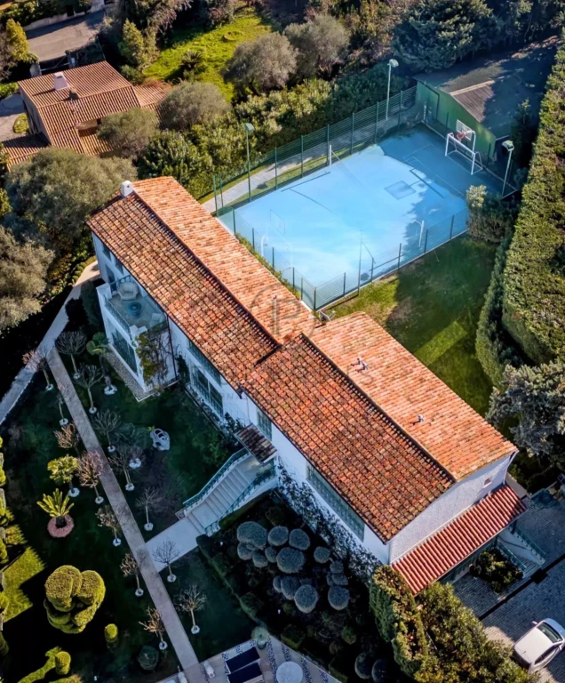Vue aérienne d'une maison avec un toit en tuiles rouges et un terrain de sport attenant, entourée de verdure.