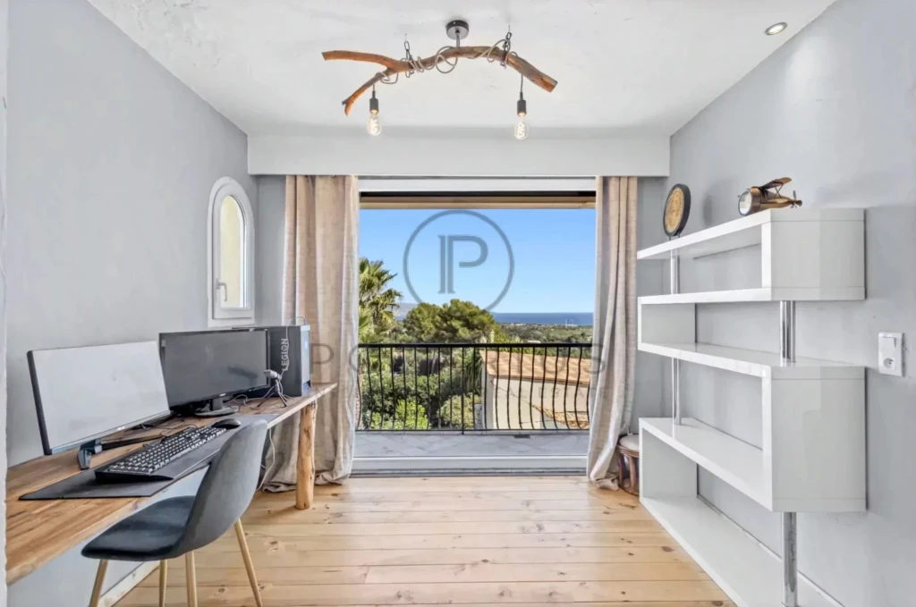 Un bureau moderne avec un bureau en bois, un ordinateur et une étagère blanche, offrant une vue sur un paysage extérieur à travers une grande fenêtre.