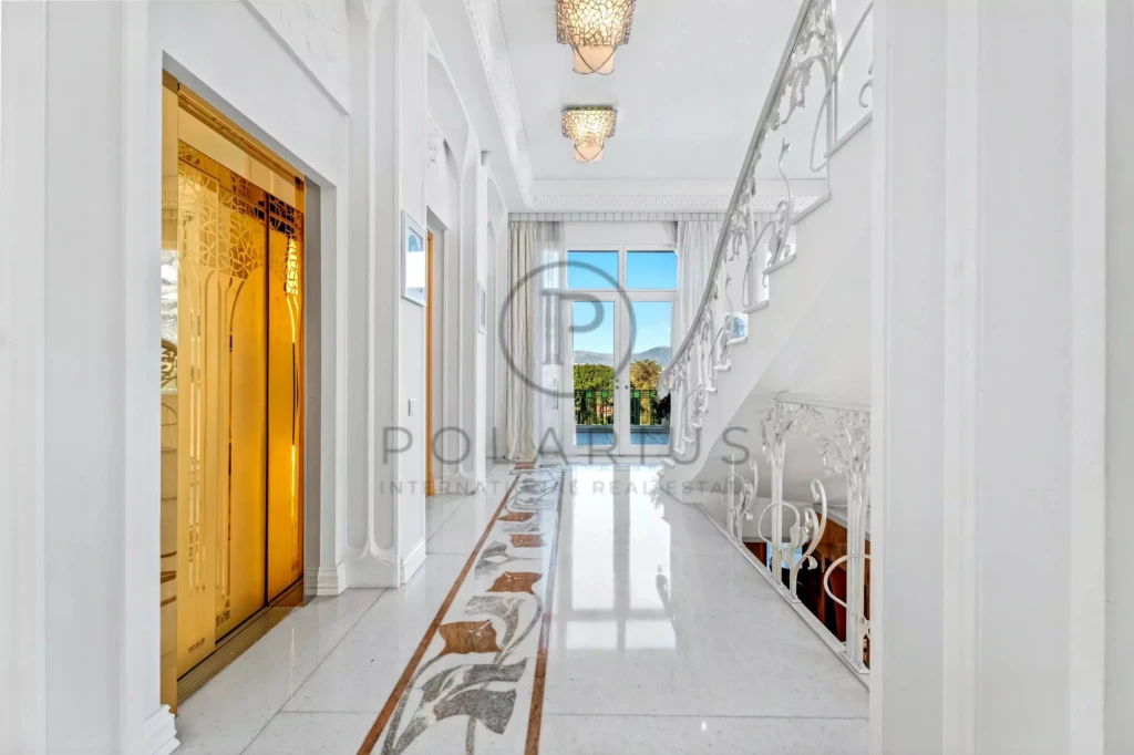 Un hall d'entrée élégant avec un sol en marbre brillant, un ascenseur doré à gauche et un escalier à droite, menant à des portes vitrées ouvertes sur une vue extérieure.