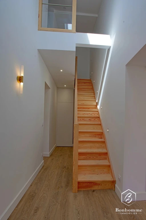 Un escalier en bois se trouve dans un intérieur moderne et lumineux, avec un sol en parquet clair et des murs blancs.