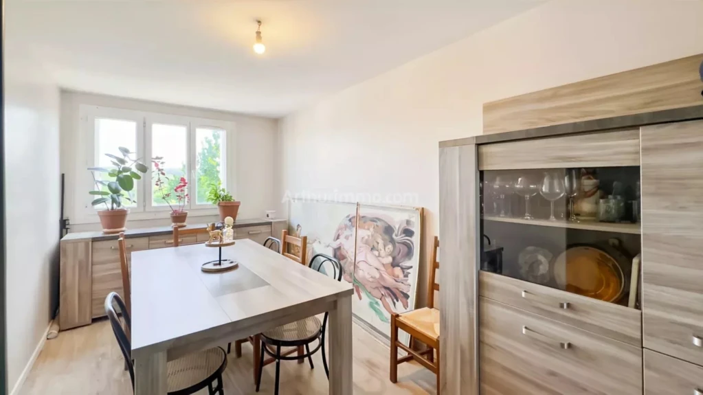 PONTHIERRY F4 Une salle à manger lumineuse avec une grande table en bois, des chaises, et des plantes près de la fenêtre. Un buffet avec des portes vitrées est visible sur la droite.
