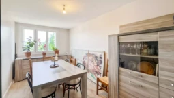 PONTHIERRY F4 Une salle à manger lumineuse avec une grande table en bois, des chaises, et des plantes près de la fenêtre. Un buffet avec des portes vitrées est visible sur la droite.