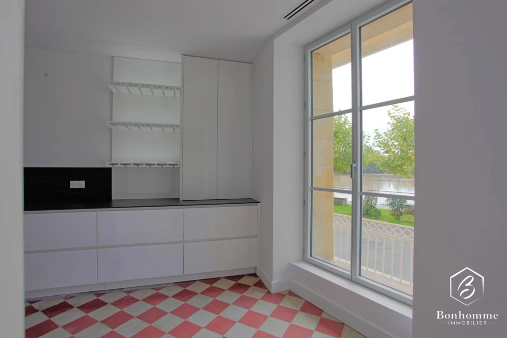 Une petite cuisine lumineuse avec des placards blancs, un sol à carreaux rouges et blancs, et une grande fenêtre donnant sur l'extérieur.