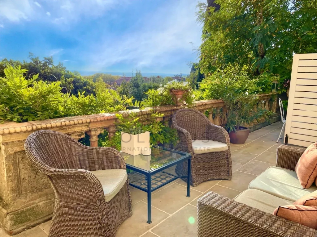 Deux fauteuils en rotin sont disposés autour d'une table en verre sur une terrasse ensoleillée, entourée de végétation luxuriante.