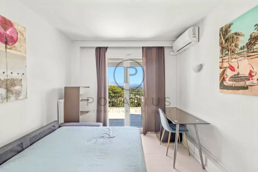 Chambre lumineuse avec un lit, un bureau et une grande fenêtre offrant une vue sur un paysage extérieur.
