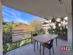 Une terrasse avec une table et deux chaises, entourée de végétation luxuriante et offrant une vue sur des arbres et des bâtiments voisins sous un ciel bleu.