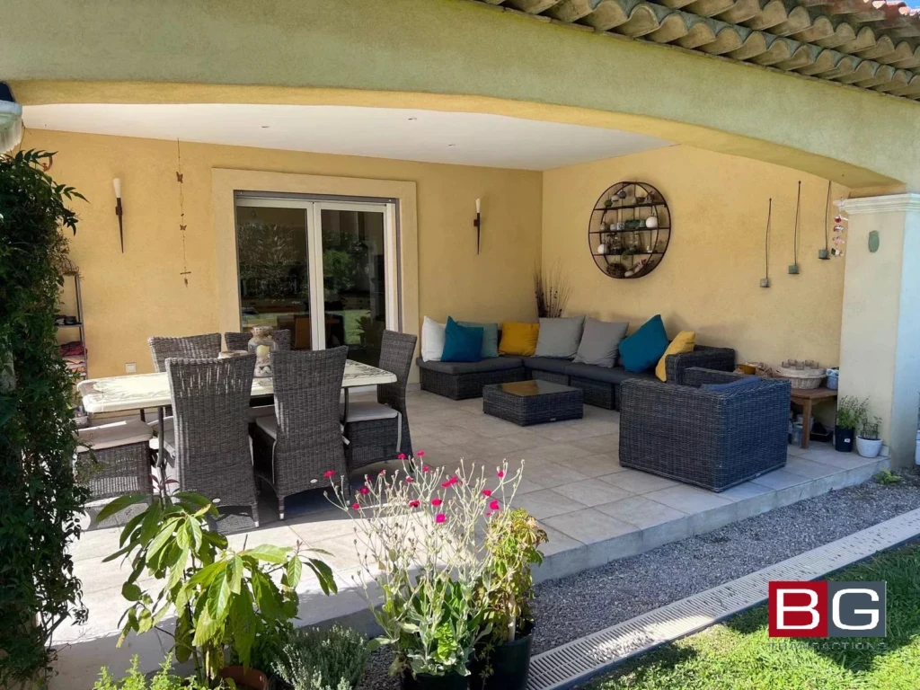 Terrasse extérieure avec table, chaises et canapé en rotin, ornée de coussins colorés et entourée de plantes.