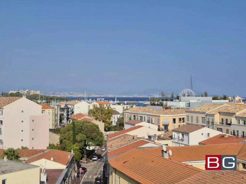 Vue urbaine avec des toits en tuiles, des bâtiments colorés, et la mer en arrière-plan sous un ciel bleu clair.
