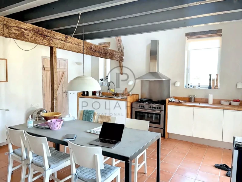 Une cuisine lumineuse avec une grande table, des chaises blanches, une hotte moderne, et des poutres apparentes.