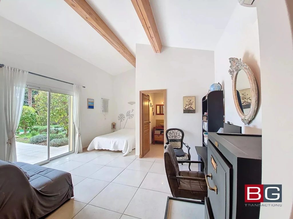 Une chambre lumineuse avec un lit simple, des poutres en bois, un grand miroir, et une porte-fenêtre donnant sur un jardin.