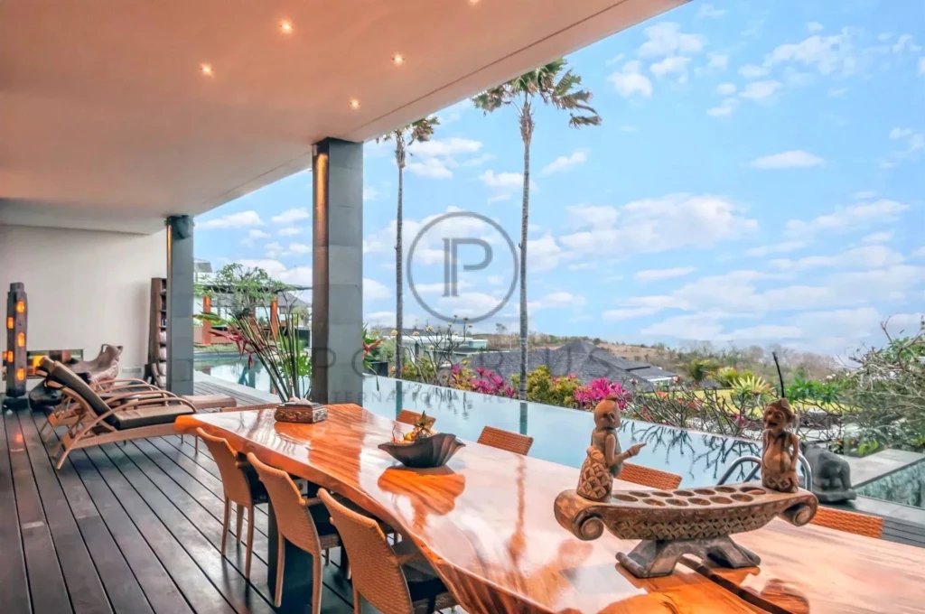 Terrasse moderne avec une longue table en bois, vue sur piscine et palmiers sous un ciel bleu.