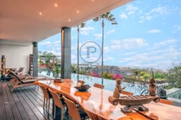 Terrasse moderne avec une longue table en bois, vue sur piscine et palmiers sous un ciel bleu.