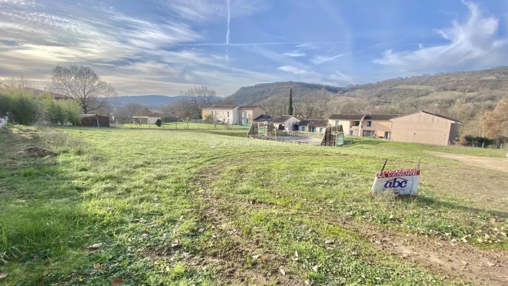 Un paysage rural avec un champ vert, des maisons au loin et des collines en arrière-plan, sous un ciel partiellement nuageux.
