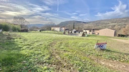 Un paysage rural avec un champ vert, des maisons au loin et des collines en arrière-plan, sous un ciel partiellement nuageux.