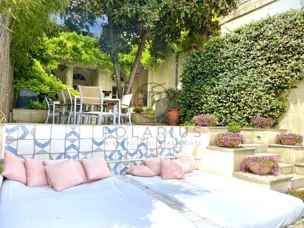 Un jardin accueillant avec des lits de repos blancs et des coussins roses, entouré par des plantes vertes et des fleurs colorées, à côté d'une table et des chaises pour manger en plein air.