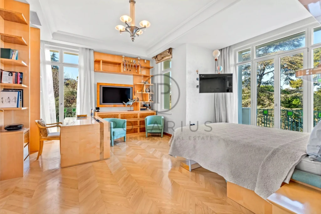 Une chambre lumineuse avec parquet en bois, un lit, un bureau, des étagères, deux fauteuils verts, et de grandes fenêtres donnant sur un jardin.