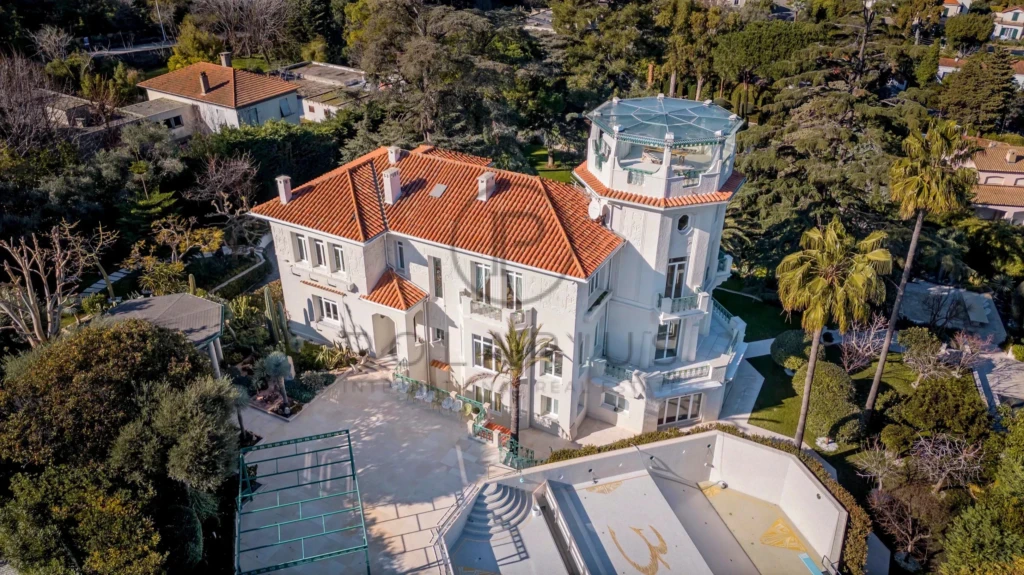 Grande villa blanche avec toit en tuiles rouges entourée de jardins et palmiers.