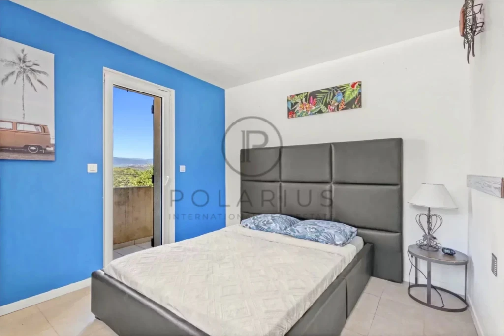 Chambre moderne avec un mur bleu, un lit double, une porte-fenêtre et décoration murale tropicale.