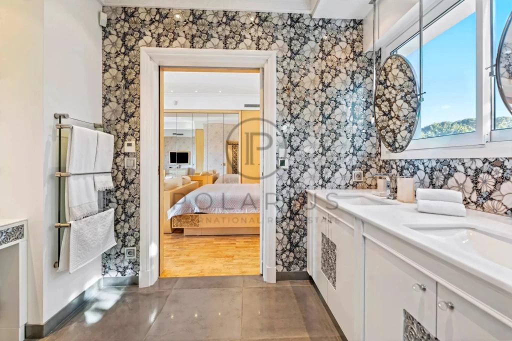 Une salle de bain élégante avec un papier peint floral, double vasque, et vue sur une chambre lumineuse.