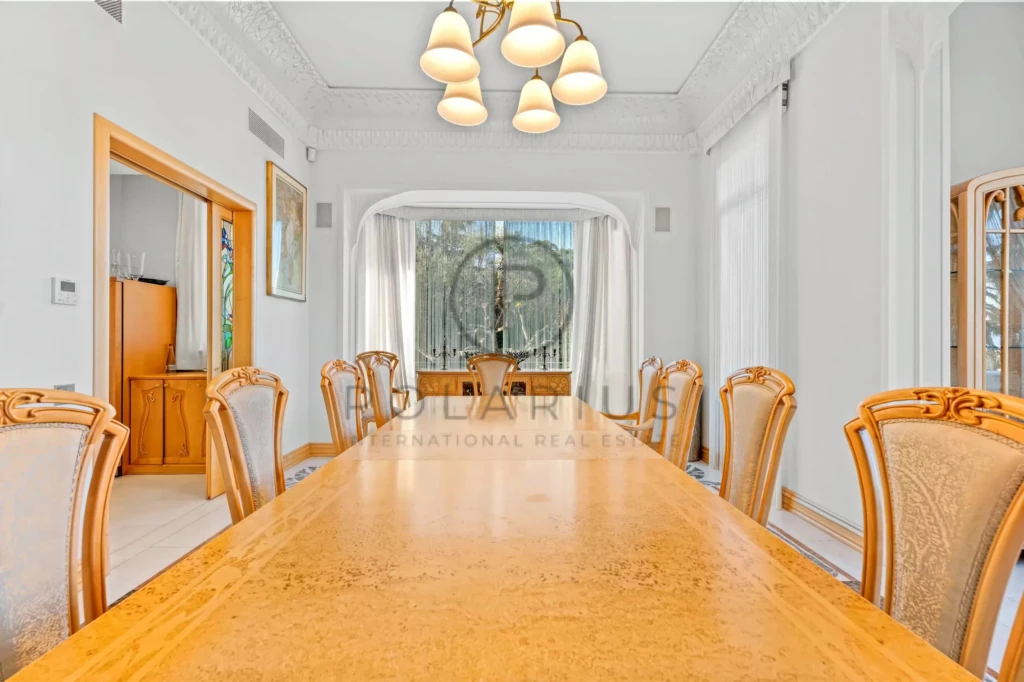 Salle à manger élégante avec une grande table en bois, des chaises ornées, un lustre et une fenêtre donnant sur le jardin.