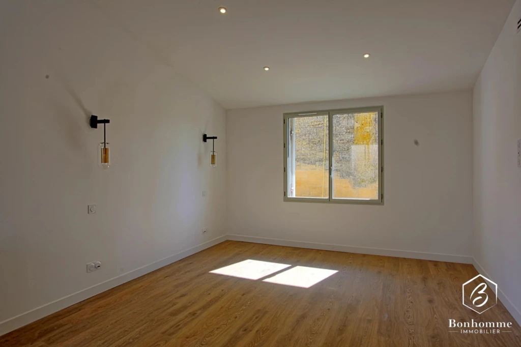 Une pièce vide avec un sol en bois, murs blancs, une fenêtre et des luminaires modernes.