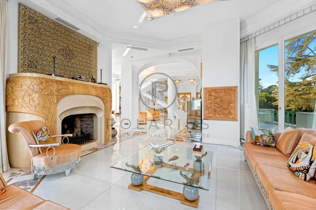 Un salon élégant avec une cheminée ornementée, des meubles en cuir beige, et une table basse en verre, baigné de lumière naturelle.