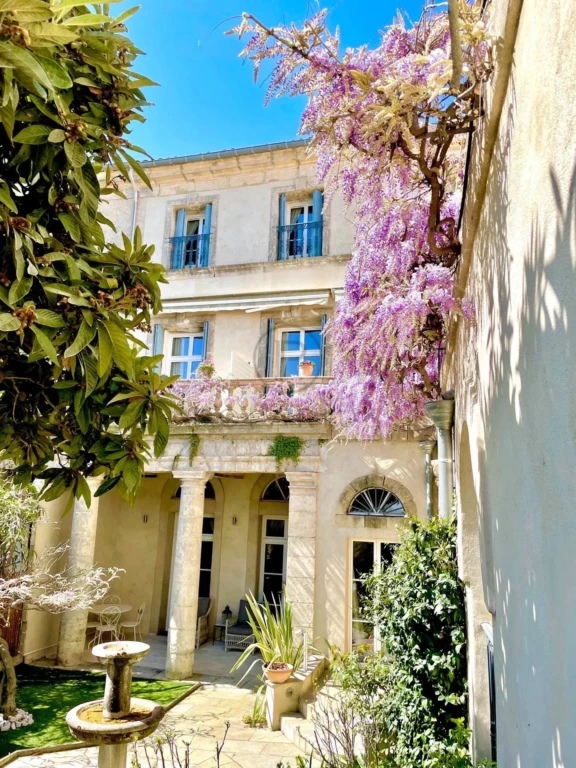 Une charmante maison ancienne avec des glycines en fleurs, entourée de verdure sous un ciel bleu.