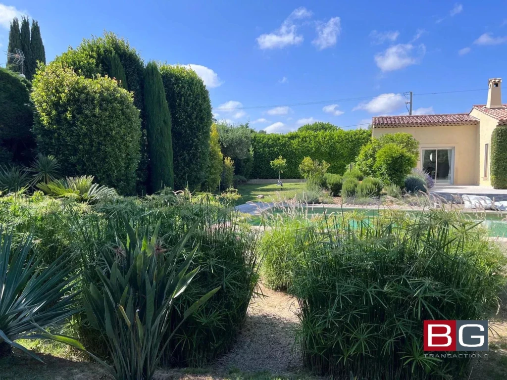 Un jardin paisible avec une piscine, entouré de verdure et d'arbustes, sous un ciel bleu clair. Une maison se trouve à droite de l'image.