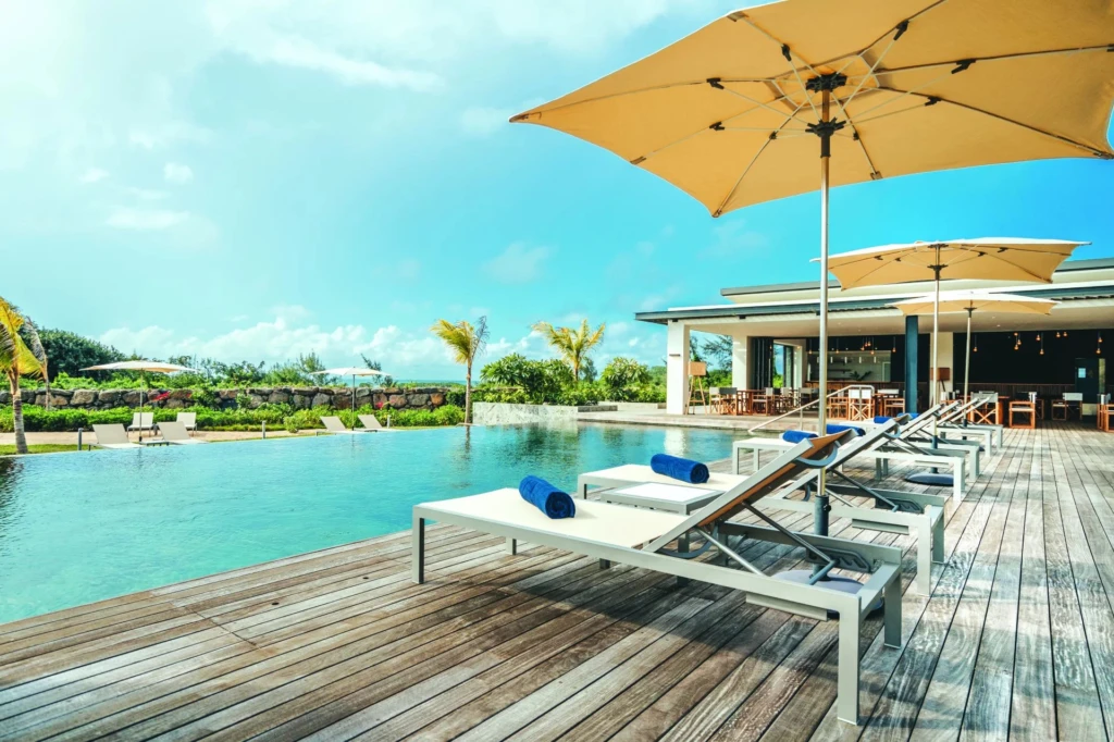 Terrasse en bois avec transats près d'une piscine, entourée de parasols sous un ciel bleu.