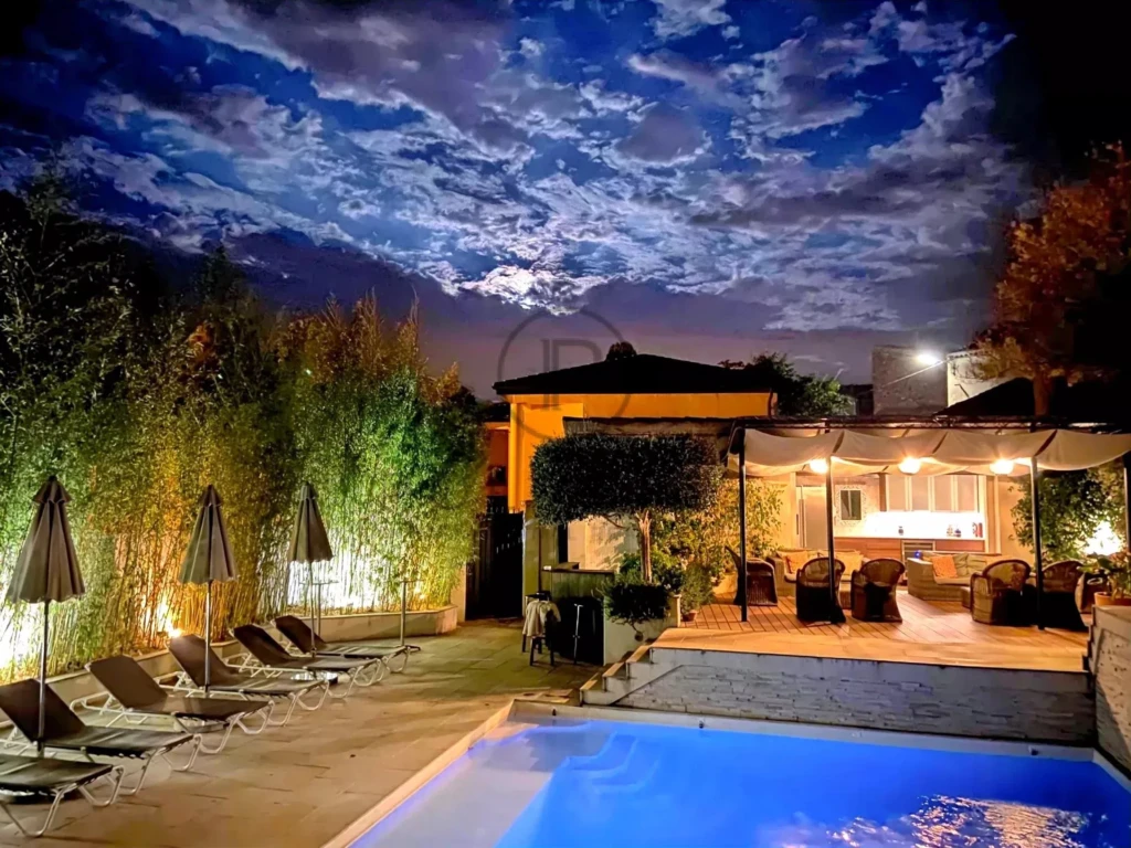 Piscine éclairée et terrasse aménagée sous un ciel nocturne nuageux.