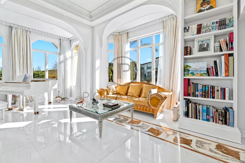 Cette photo montre un salon lumineux et élégant avec un canapé doré, un piano blanc et une bibliothèque remplie de livres.